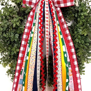 May include: A decorative wreath featuring a large red and white gingham bow with long, cascading ribbons in various patterns, including school-themed prints with apples, pencils, crayons, alphabet letters, and solid yellow, green, and navy blue.