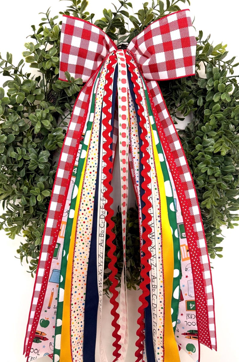 May include: A decorative wreath featuring a large red and white gingham bow with long, cascading ribbons in various patterns, including school-themed prints with apples, pencils, crayons, alphabet letters, and solid yellow, green, and navy blue.