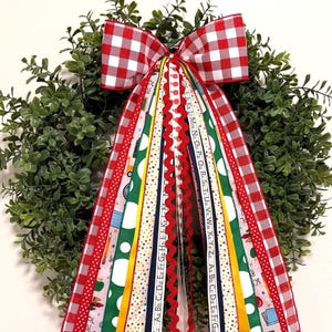 May include: A green boxwood wreath adorned with a large red and white gingham bow and multiple hanging ribbons. The ribbons feature school-themed patterns including alphabets, crayons, globes, and books in red, green, yellow, and blue colors.