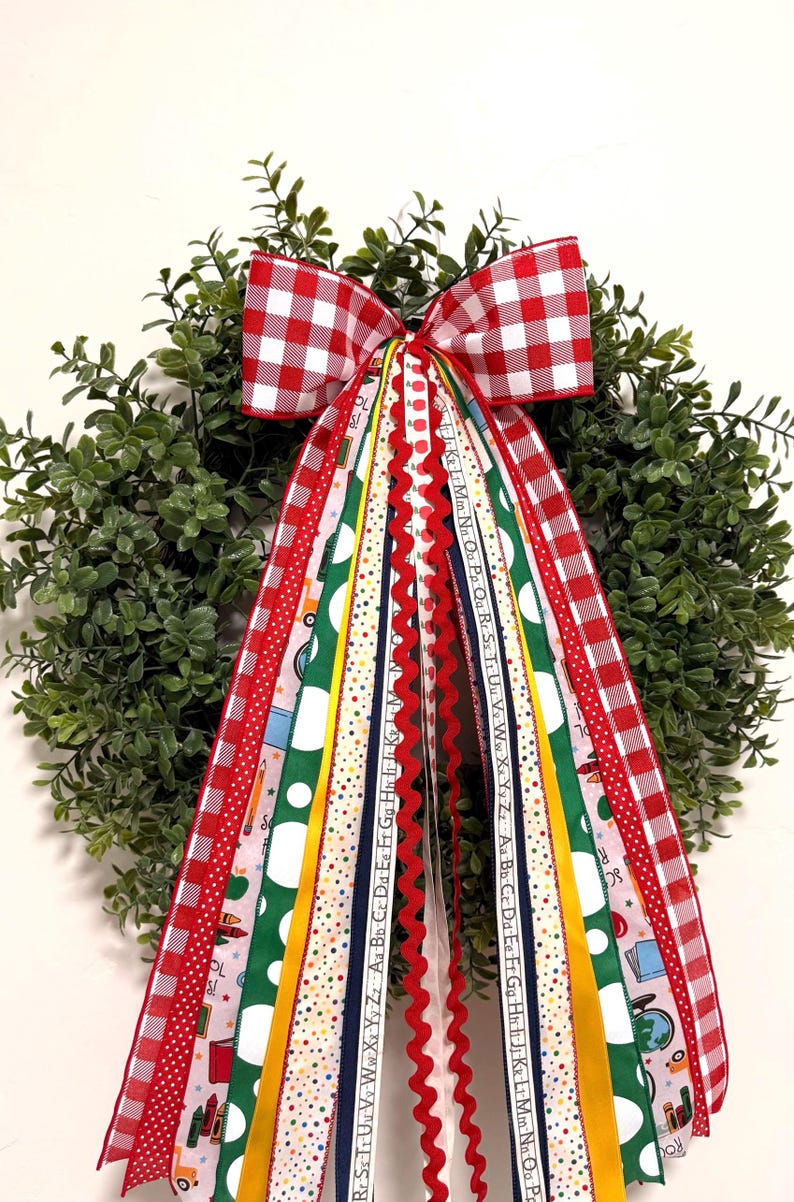 May include: A green boxwood wreath adorned with a large red and white gingham bow and multiple hanging ribbons. The ribbons feature school-themed patterns including alphabets, crayons, globes, and books in red, green, yellow, and blue colors.