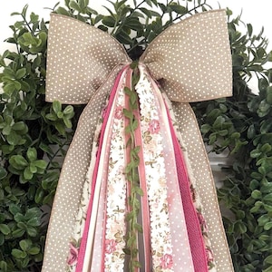 May include: A decorative wreath bow with a large tan polka-dot bow and cascading ribbons. The ribbons include floral patterns, solid pink, and white with green vine accents. The wreath is surrounded by green foliage.