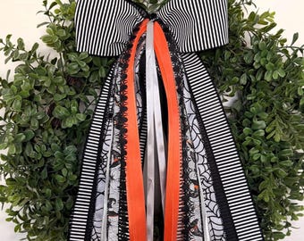 May include: A decorative bow featuring black and white striped ribbon, accented with long streamers of orange satin, solid black ribbon, and white ribbon printed with black spiderwebs and witch hat patterns, all layered over a green leaf wreath.