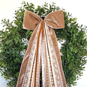 May include: A decorative bow made of tan velvet fabric with long, cascading ribbons in various patterns including white leaf prints, brown and white gingham, solid tan, and textured jute, centered over a circular green faux boxwood wreath.