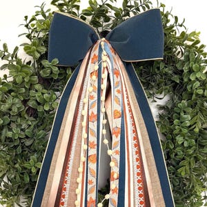 May include: A decorative wreath featuring a large navy blue velvet bow with gold trim, accented by long, cascading ribbons in shades of navy, brown, tan, and cream, including a patterned ribbon with orange autumn leaves and a string of small cream pom-poms.
