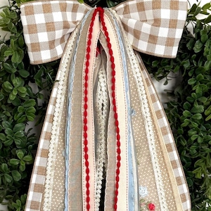 May include: A decorative wreath with a large bow. The bow features a brown and white gingham pattern and multiple ribbons in various colors and textures, including red, beige, and blue. The wreath is surrounded by green foliage.