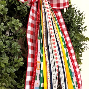 May include: A decorative wreath bow featuring a red and white gingham pattern with long, cascading ribbons. The ribbons display school-themed motifs including apples, pencils, and the alphabet in various colors like green, yellow, and blue.