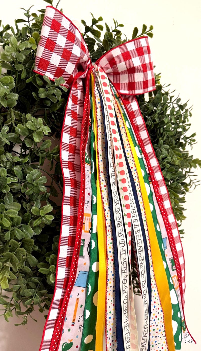 May include: A decorative wreath bow featuring a red and white gingham pattern with long, cascading ribbons. The ribbons display school-themed motifs including apples, pencils, and the alphabet in various colors like green, yellow, and blue.