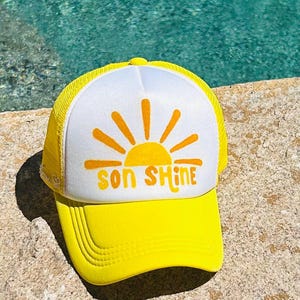 May include: Yellow and white trucker hat with the words "SON SHINE" in orange, along with a sun graphic. The hat is sitting on a stone surface, with a pool of water in the background.