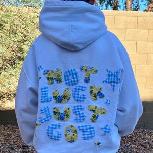 May include: White hoodie with the text "NOT LUCK JUST GOT" appliqued on the back in blue gingham and yellow floral fabric. The hoodie has a drawstring hood and is worn with denim shorts.