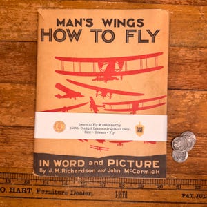 May include: A vintage book titled "Man's Wings How to Fly" with red airplane illustrations on a tan cover. The book includes text about 1930s cockpit lessons and Quaker Oats. A white band with text wraps around the book.