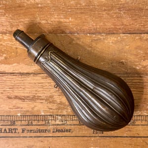 May include: An antique bronze powder flask with a ribbed design. The flask features a cylindrical spout and decorative detailing near the top. Two small loops are attached to the side. It rests on a wooden surface.