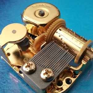 Op de afbeelding: Een goudkleurig muziekdoosmechanisme met een opwindsleutel, een kam en een metalen plaat met een reeks pinnen. Het mechanisme is gemarkeerd met "Sankyo K220".