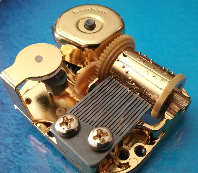 Op de afbeelding: Een goudkleurig muziekdoosmechanisme met een opwindsleutel, een kam en een metalen plaat met een reeks pinnen. Het mechanisme is gemarkeerd met "Sankyo K220".