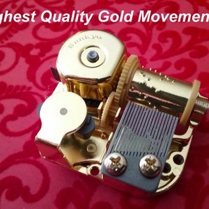 May include: Gold-colored music box movement with a winding key, a comb, and a small metal plate with two screws. The movement is on a red background.