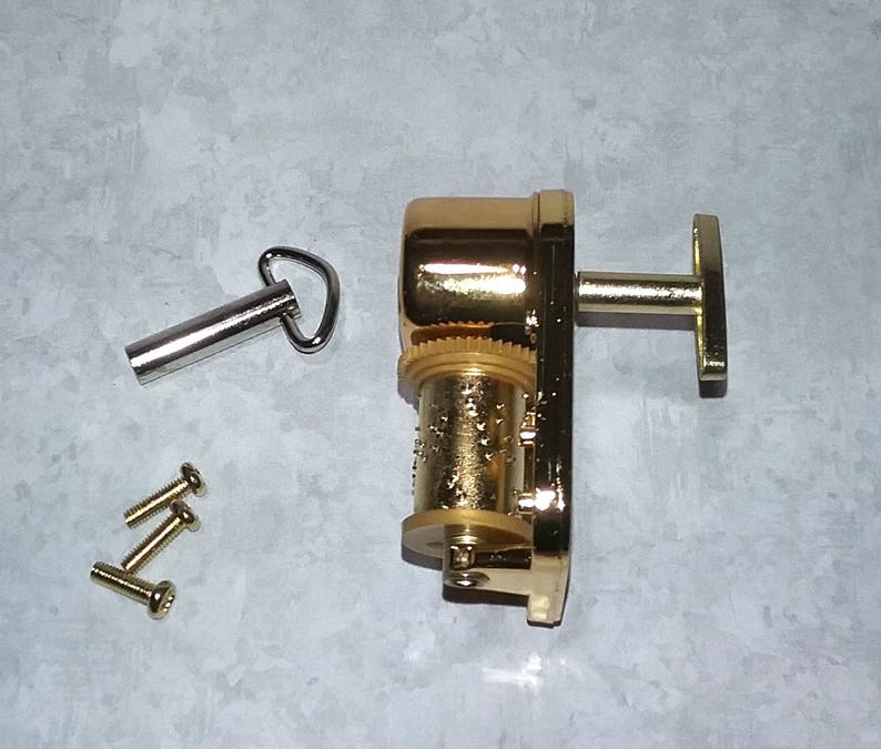 May include: A gold-colored music box mechanism with a winding key and a T-shaped handle. The mechanism is accompanied by a silver-colored winding key and four small gold-colored screws.