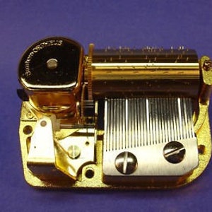 May include: A gold-colored music box mechanism with a winding key and a metal comb. The mechanism is designed to play a tune when wound.