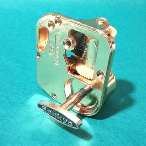 Op de afbeelding: Een goudkleurig muziekdoosmechanisme met een opwindsleutel. Het mechanisme heeft zichtbare tandwielen en een Sankyo-label op de sleutel.
