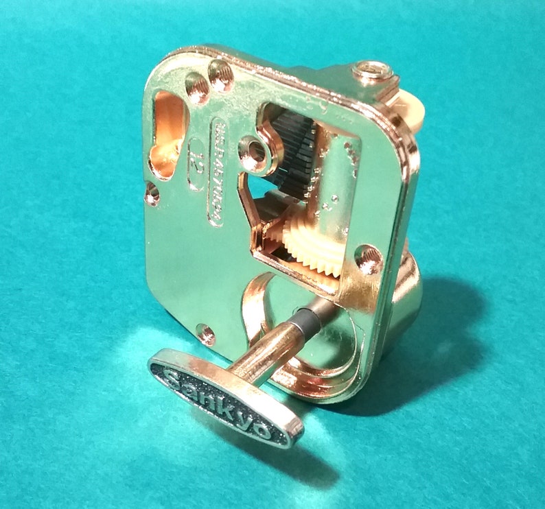 Op de afbeelding: Een goudkleurig muziekdoosmechanisme met een opwindsleutel. Het mechanisme heeft zichtbare tandwielen en een Sankyo-label op de sleutel.