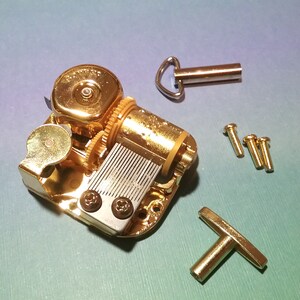 May include: Gold-toned music box mechanism with a winding key, a T-shaped crank, and four screws. The mechanism has a silver-colored comb and a gold-toned cylinder.