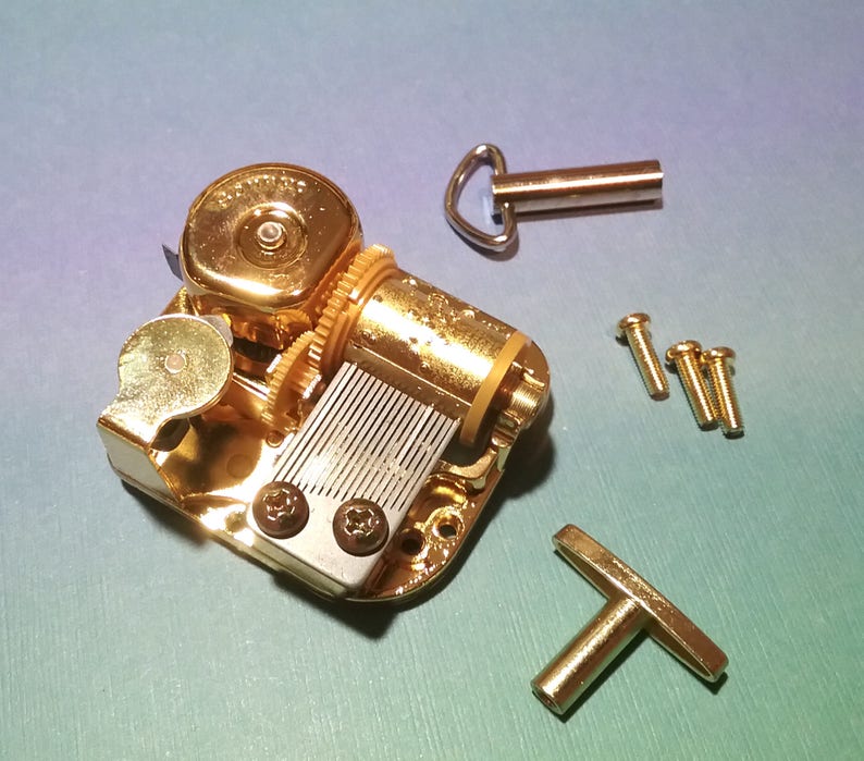 May include: Gold-toned music box mechanism with a winding key, a T-shaped crank, and four screws. The mechanism has a silver-colored comb and a gold-toned cylinder.