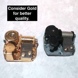 May include: Two music boxes, one gold and one silver, with a black winding key. The gold music box has a gold-colored winding key. The silver music box has a black winding key. The text "Consider Gold for better quality." is visible in the image.