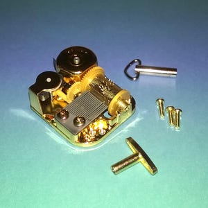 May include: A gold-colored music box mechanism with a winding key and a small metal crank. The mechanism is on a blue surface with four small screws.