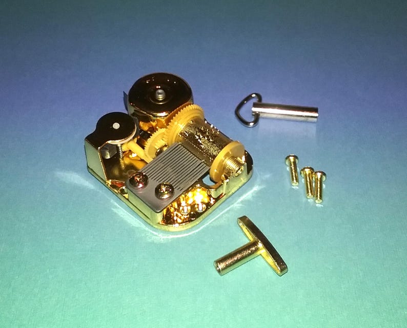 May include: A gold-colored music box mechanism with a winding key and a small metal crank. The mechanism is on a blue surface with four small screws.
