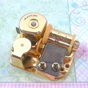Op de afbeelding: Gouden muziekdoosmechanisme met een opwindsleutel en een metalen kam. Het mechanisme is gemarkeerd met "SANKYO".
