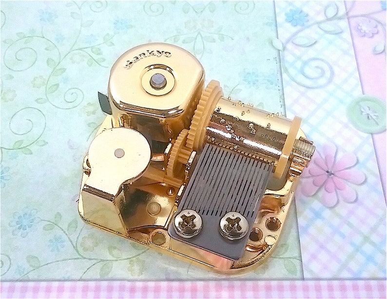 Op de afbeelding: Gouden muziekdoosmechanisme met een opwindsleutel en een metalen kam. Het mechanisme is gemarkeerd met "SANKYO".