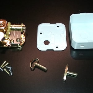 Op de afbeelding: Een goudkleurig muziekdoosmechanisme met een witte plastic doos, een opwindsleutel en een kleine metalen staaf. De muziekdoos is gedemonteerd en ligt op een zwart oppervlak.