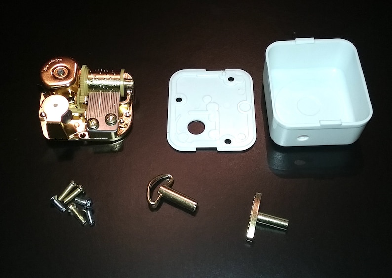Op de afbeelding: Een goudkleurig muziekdoosmechanisme met een witte plastic doos, een opwindsleutel en een kleine metalen staaf. De muziekdoos is gedemonteerd en ligt op een zwart oppervlak.