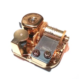 May include: Gold-toned music box mechanism with a winding key, a metal comb, and two screws. The mechanism is designed to play a tune when wound.