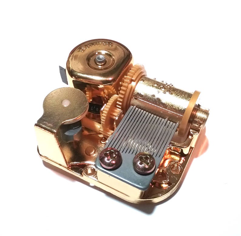May include: Gold-toned music box mechanism with a winding key, a metal comb, and two screws. The mechanism is designed to play a tune when wound.