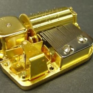 May include: A gold-colored music box mechanism with a winding key and a small, round, gold-colored music box. The mechanism has a series of gears and a metal comb.