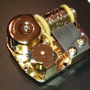 Op de afbeelding: Een goudkleurig muziekdoosmechanisme met een opwindsleutel en een kam van metalen tanden. Het mechanisme is van metaal en heeft een gepolijste afwerking.