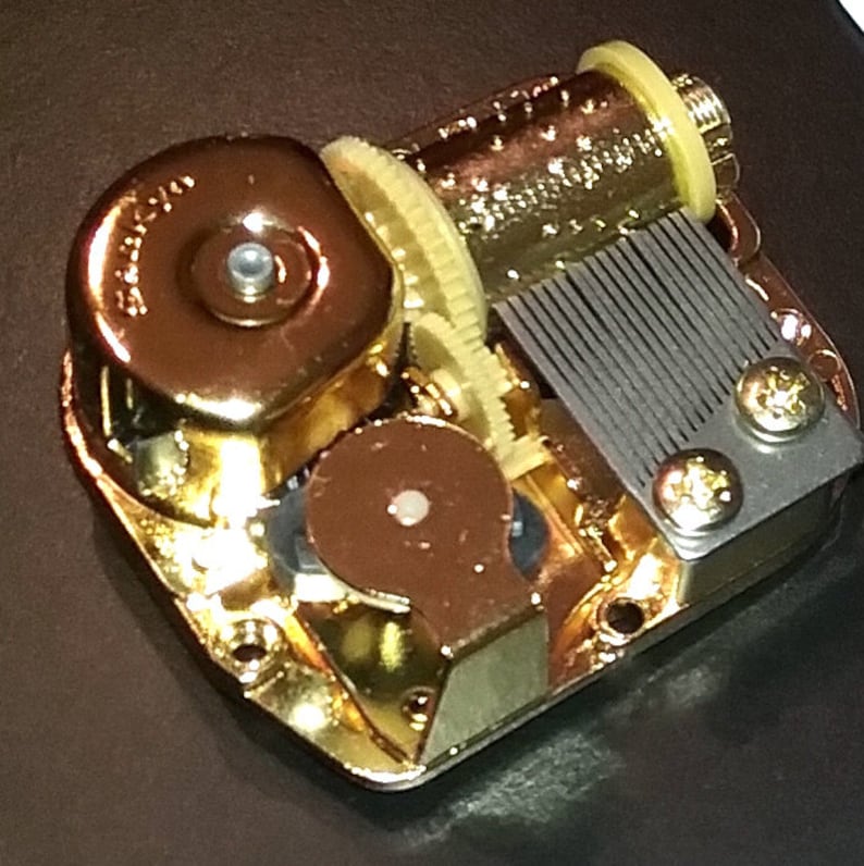Op de afbeelding: Een goudkleurig muziekdoosmechanisme met een opwindsleutel en een kam van metalen tanden. Het mechanisme is van metaal en heeft een gepolijste afwerking.