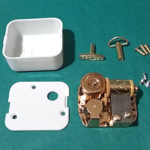 Op de afbeelding: Een goudkleurig muziekdoosmechanisme met een witte kunststof behuizing, een opwindsleutel en schroeven. De muziekdoos is gedemonteerd en klaar om in elkaar te worden gezet.