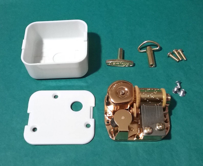 Op de afbeelding: Een goudkleurig muziekdoosmechanisme met een witte kunststof behuizing, een opwindsleutel en schroeven. De muziekdoos is gedemonteerd en klaar om in elkaar te worden gezet.