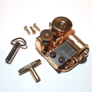 May include: Gold-toned music box mechanism with a winding key and four small screws. The mechanism has a gray metal plate with a series of holes and a winding key with a loop on one end and a T-shaped handle on the other.