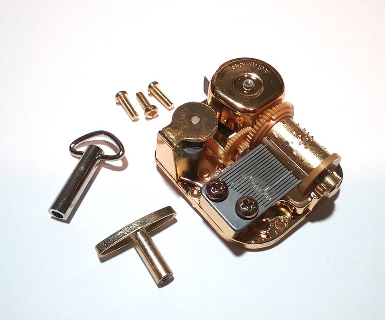 May include: Gold-toned music box mechanism with a winding key and four small screws. The mechanism has a gray metal plate with a series of holes and a winding key with a loop on one end and a T-shaped handle on the other.
