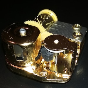 Op de afbeelding: Een goudkleurig muziekdoosmechanisme met een opwindsleutel, tandwielen en een metalen kam. Het mechanisme is ontworpen om muziek af te spelen wanneer het wordt opgewonden.