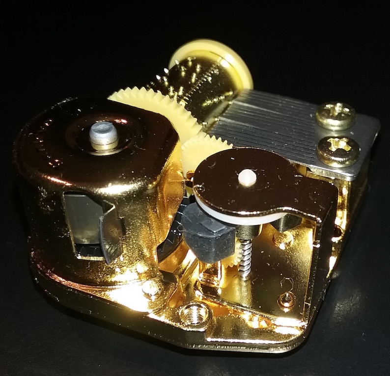 Op de afbeelding: Een goudkleurig muziekdoosmechanisme met een opwindsleutel, tandwielen en een metalen kam. Het mechanisme is ontworpen om muziek af te spelen wanneer het wordt opgewonden.