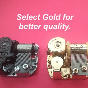 Op de afbeelding: Twee muziekdoosmechanismen, een zwart en een gouden. De tekst "Select Gold for better quality." wordt boven de mechanismen weergegeven.