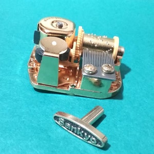 Op de afbeelding: Gouden muziekdoosmechanisme met een opwindsleutel en een kruk. Het mechanisme bevindt zich op een blauwe ondergrond. De kruk is gemarkeerd met "Sankyo".
