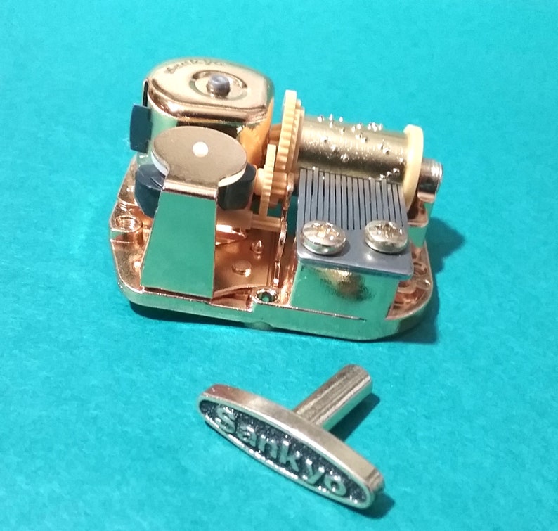 Op de afbeelding: Gouden muziekdoosmechanisme met een opwindsleutel en een kruk. Het mechanisme bevindt zich op een blauwe ondergrond. De kruk is gemarkeerd met "Sankyo".