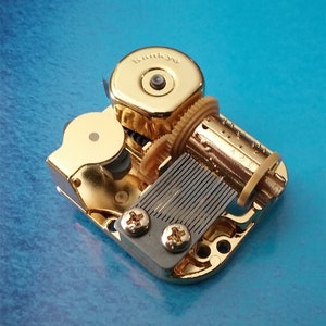 May include: A gold-colored music box mechanism with a winding key and a metal comb. The mechanism is labeled "Sunkyio".