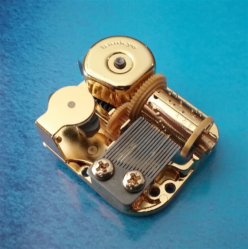 May include: A gold-colored music box mechanism with a winding key and a metal comb. The mechanism is labeled "Sunkyio".
