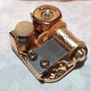May include: Gold-colored music box mechanism with a gray comb and a winding key. The mechanism has a series of gears and a small metal knob.