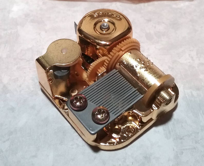 May include: Gold-colored music box mechanism with a gray comb and a winding key. The mechanism has a series of gears and a small metal knob.