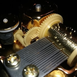 Op de afbeelding: Close-up van een goudkleurig muziekdoosmechanisme met een zwart-wit gestreepte kam en een reeks tandwielen. Het mechanisme is gemaakt van metaal en heeft een gepolijste afwerking.